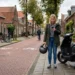 Jongere met scooter en helm op Nederlandse straat kijkt op telefoon naast geparkeerde scooter